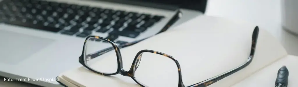 Eine Brille auf einem Notizbuch neben einem Laptop – ein Symbol für die Arbeit einer Texterin oder eines Texters in einem Unternehmen.